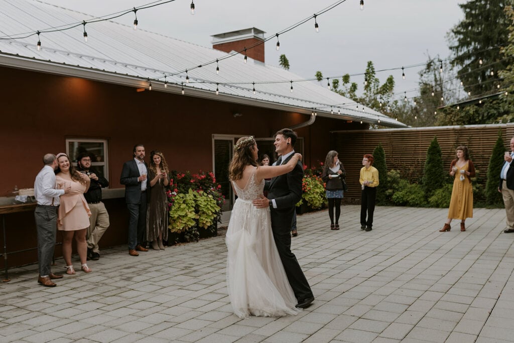 A couple stands in an outdoor courtyard dancing under string lights and surrounded by family during their elopement.