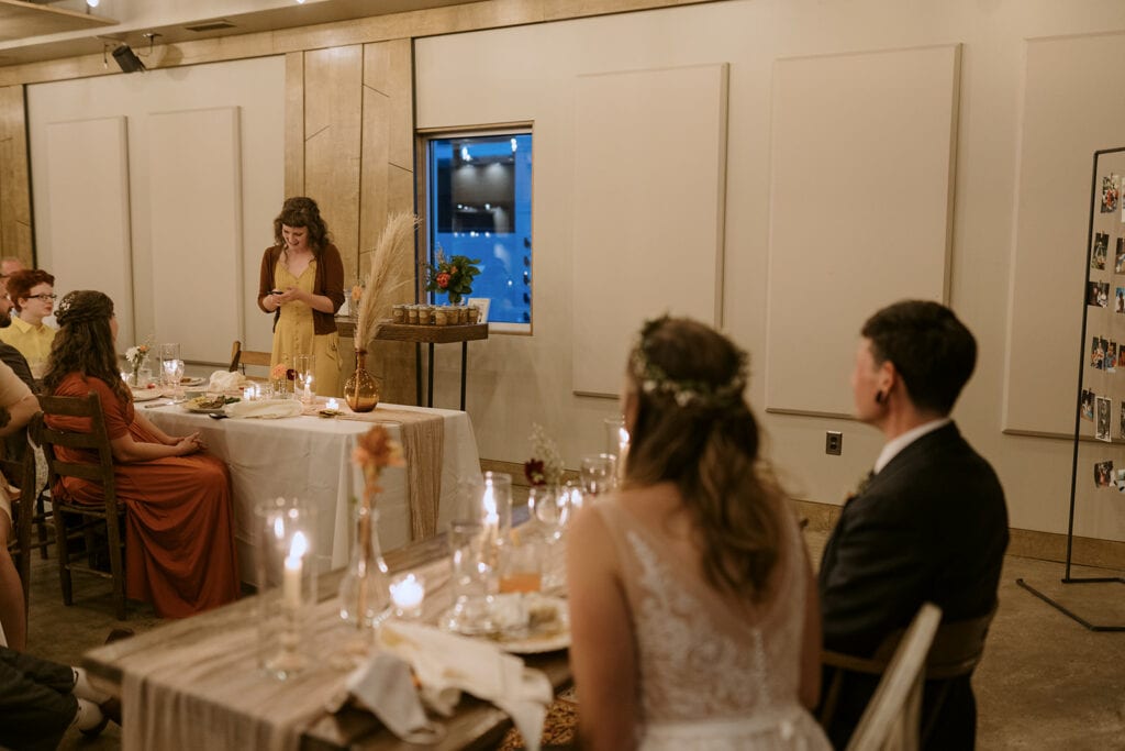 A couple watches a family member give a toast in their elopement reception hall with lit candles and low lighting.