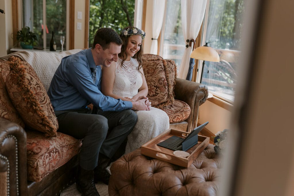 A couple in their wedding clothes sits on a couch and holds hands while smiling at the screen of a computer.