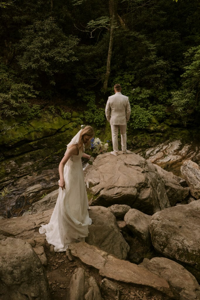 bride fixing wedding dress as she walks towards groom during their elopement on big rocks by a river in the woods.