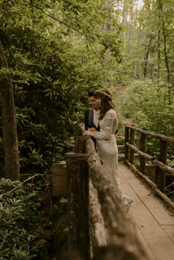 Groom standing next to bride in her Elopement Wedding Dress. on a bridge in the woods.