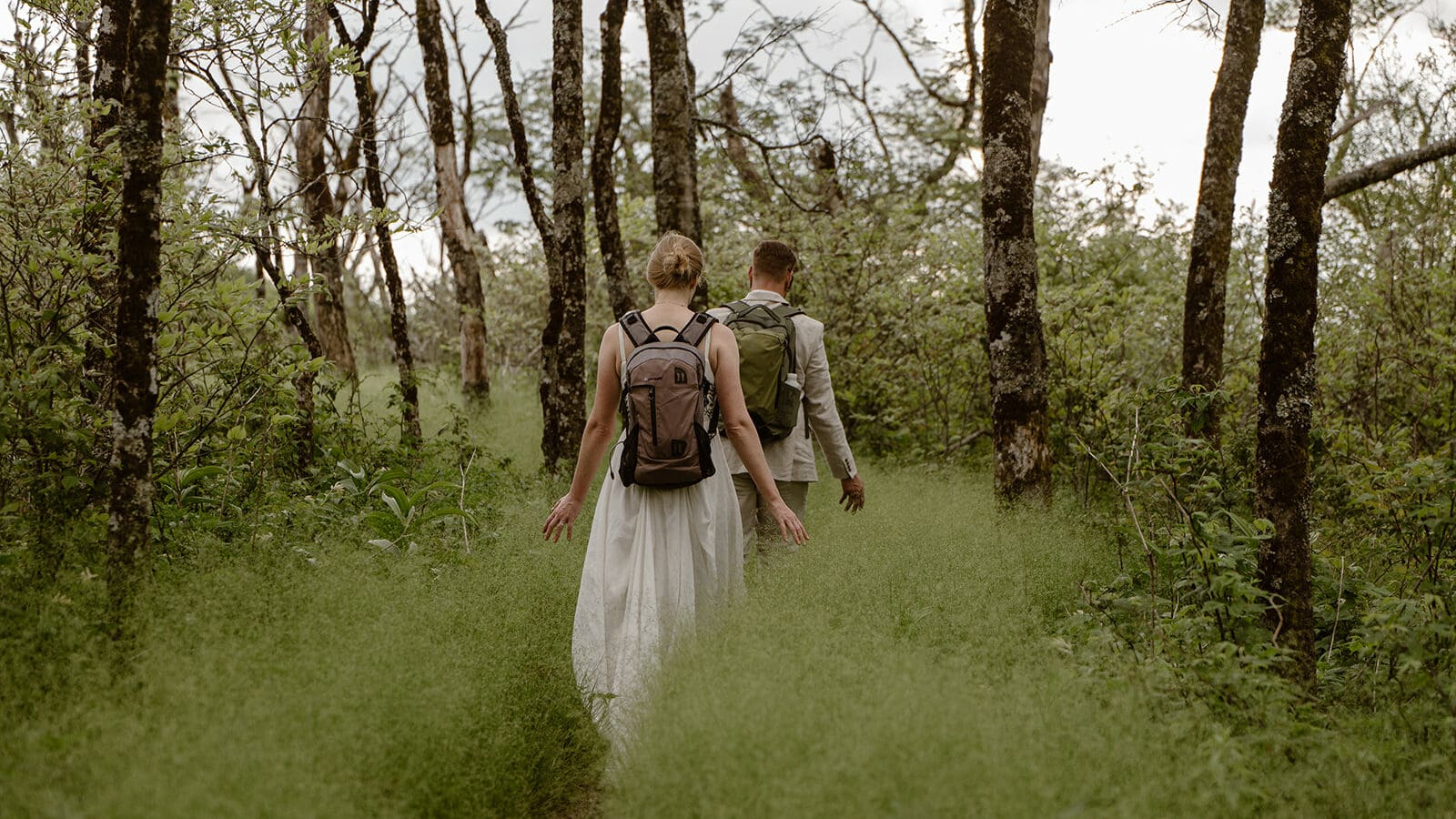 A couple is hiking through a high grassy trail in their wedding clothes in NC; one of the best places to elope.