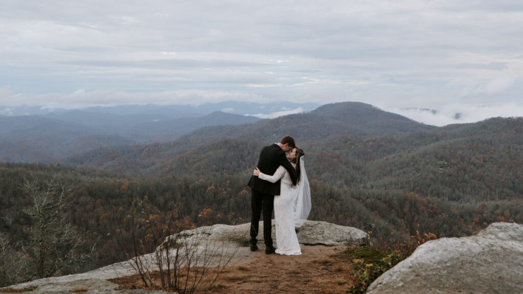 A couple is standing side by side but also face to face in clothes they eloped in on a mountain in NC. The fall colors are on the trees in the landscape and there are some cloud inversions.