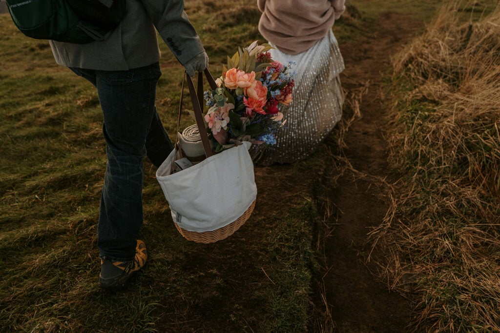 A close up of a person holding a bag full of flowers and a blanket. The other person walking with them is in a pink jacket and white sparkly dress.