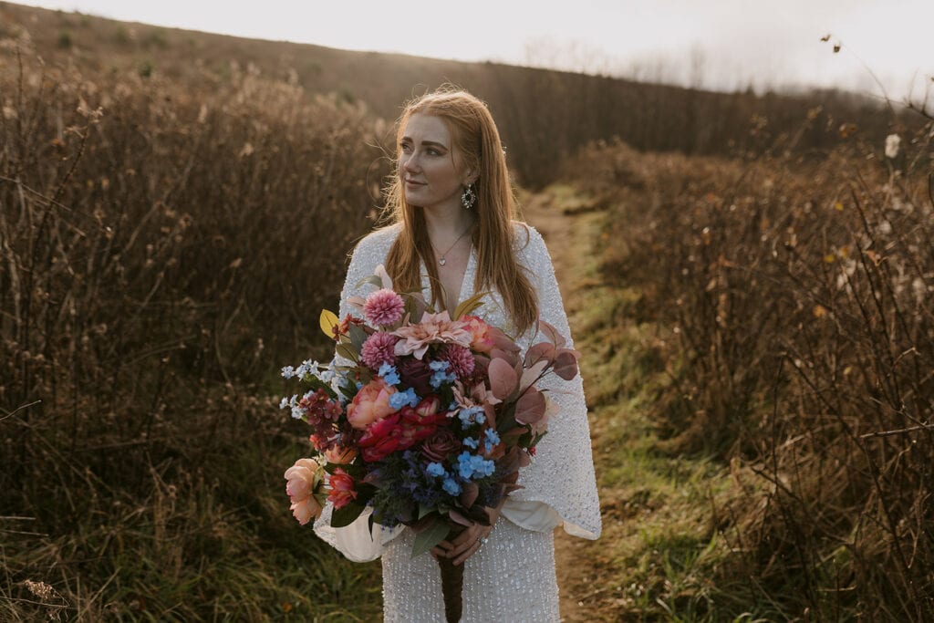 A bride is standing holding her bouquet while in her elopement dress on a trail at Max Patch.