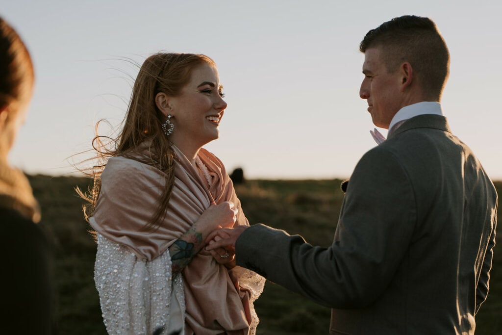 A couple is standing on a ridge for their Max patch elopement during the ceremony at sunrise. The bride is smiling at the groom and wearing a pink shawl over her dress.