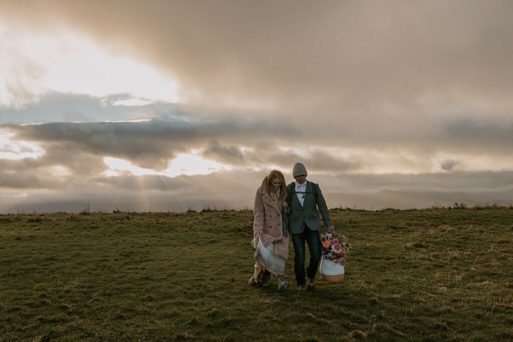 A couple walks together on a grassy area with their elopement clothes on and layers like a jacket and hat. The sun is beaming through the clouds.