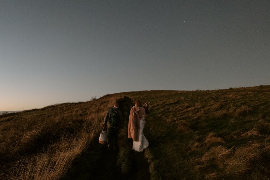 Two people are hiking up a trail at Max Patch in their elopement clothes before dawn.