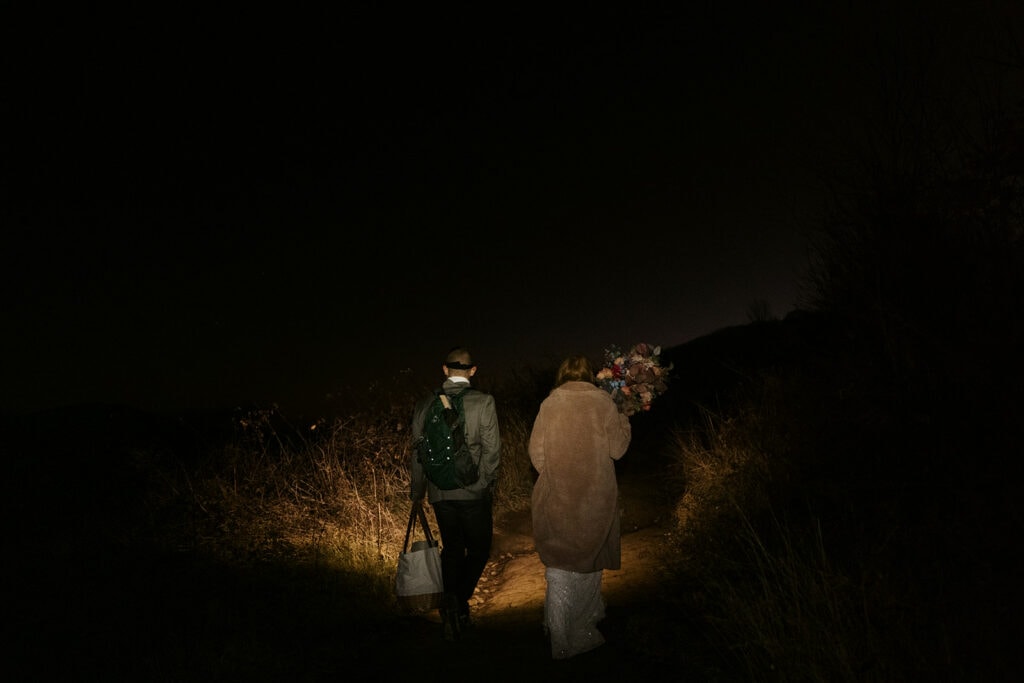 A couple is illuminated by headlamps as they hike up a dark trail in their wedding clothes.