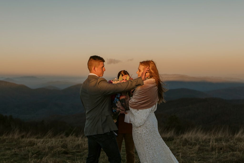 A couple is standing on a ridge for their Max patch elopement during the ceremony at sunrise. The groom is pushing the hair out of his brides face.