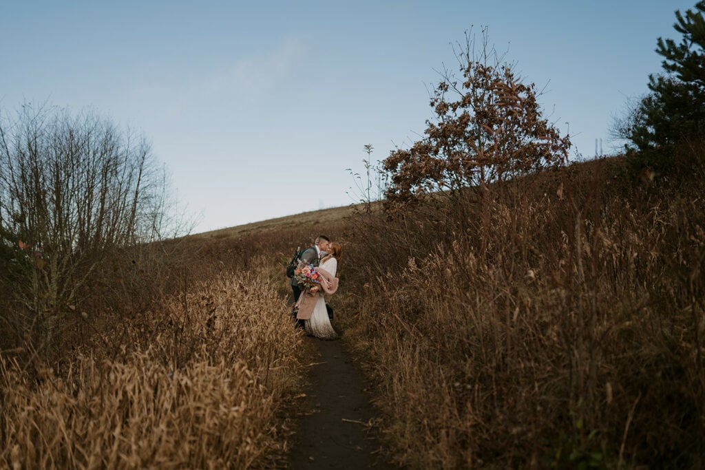 A couple is kissing in the trail at max Patch during their elopement. The trail is lined with high dried and golden grasses.