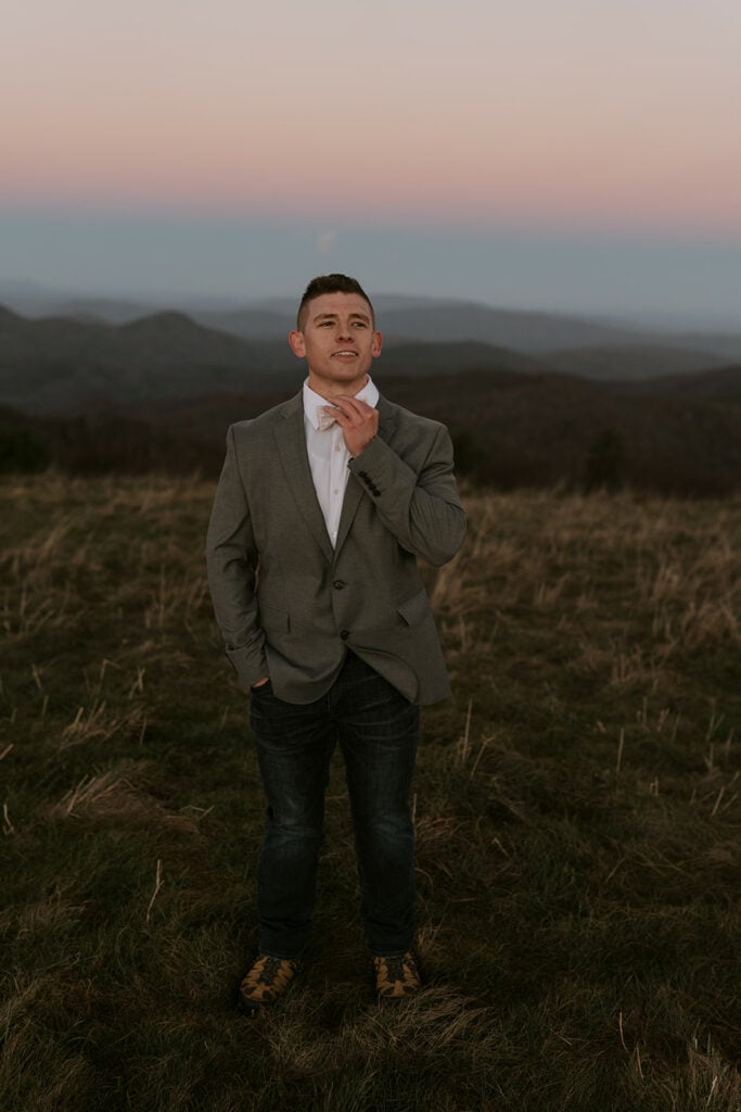 A groom is adjusting his bowtie in front of a beautiful mountain sunrise at Max Patch before his elopement.