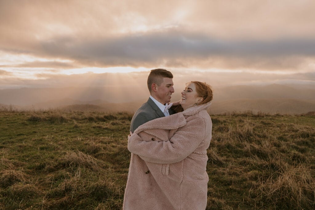 A bride is keeping her groom warm in her big pink jacket on a mountaintop during a very pink sunrise. They are smiling and laughing with each other.