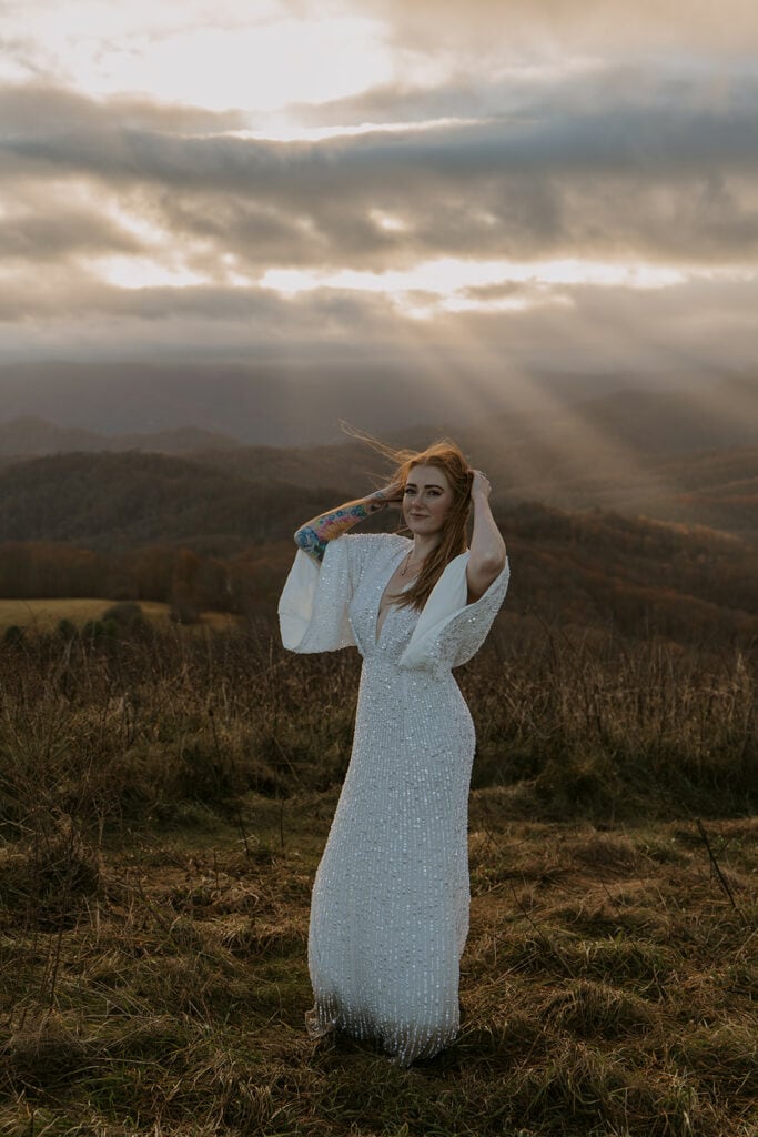A bride stands in front of clouds bursting with sunlight smiling, and holds her hair out of her eyes.