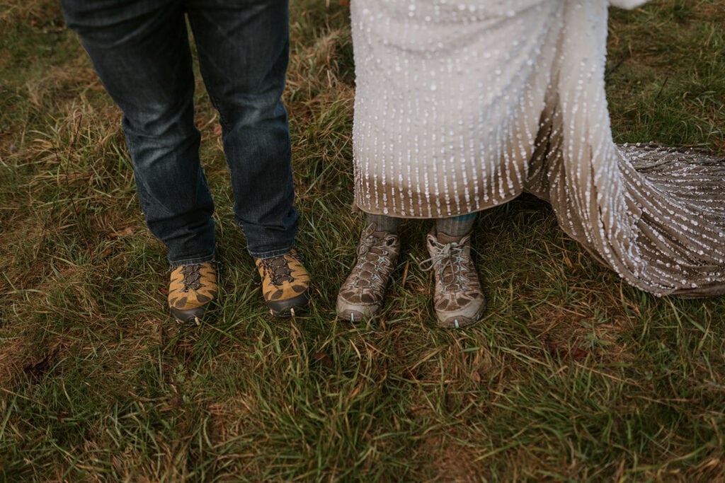 two people show off their hiking shoes. One is in a pair of jeans and the other is in a white sparkly wedding dress with the bottom all muddy