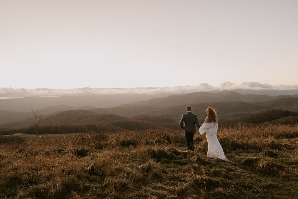 A couple is standing side by side and holding hands on a ridge for their Max patch elopement as the sun is rising and making everything glow golden.