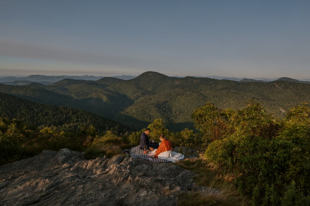 A couple is sitting on a picnic blanket on a mountaintop with a big view during their adventure elopement. The groom has a blue suit and the bride a white dress with orange shawl.