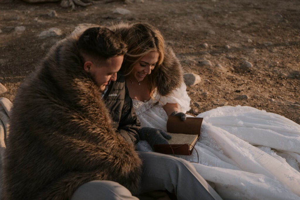 A couple is sitting and reading notes from their family during their adventure elopement. They are on the ground and wrapped in a big faux fur blanket.