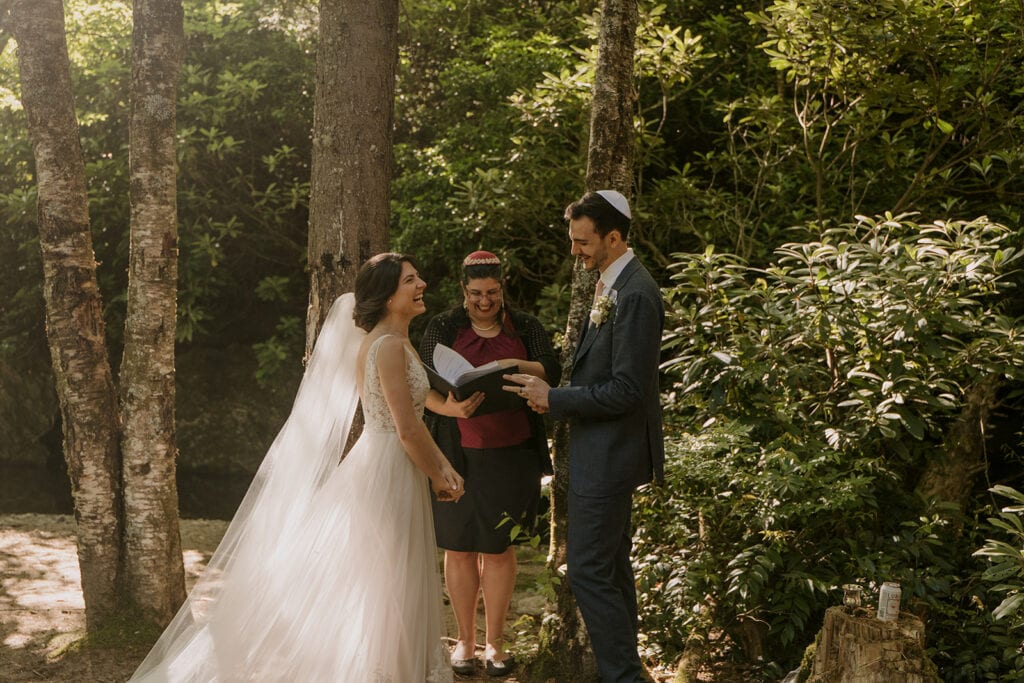 A couple stands laughing during their Jewish elopement ceremony in the forest.