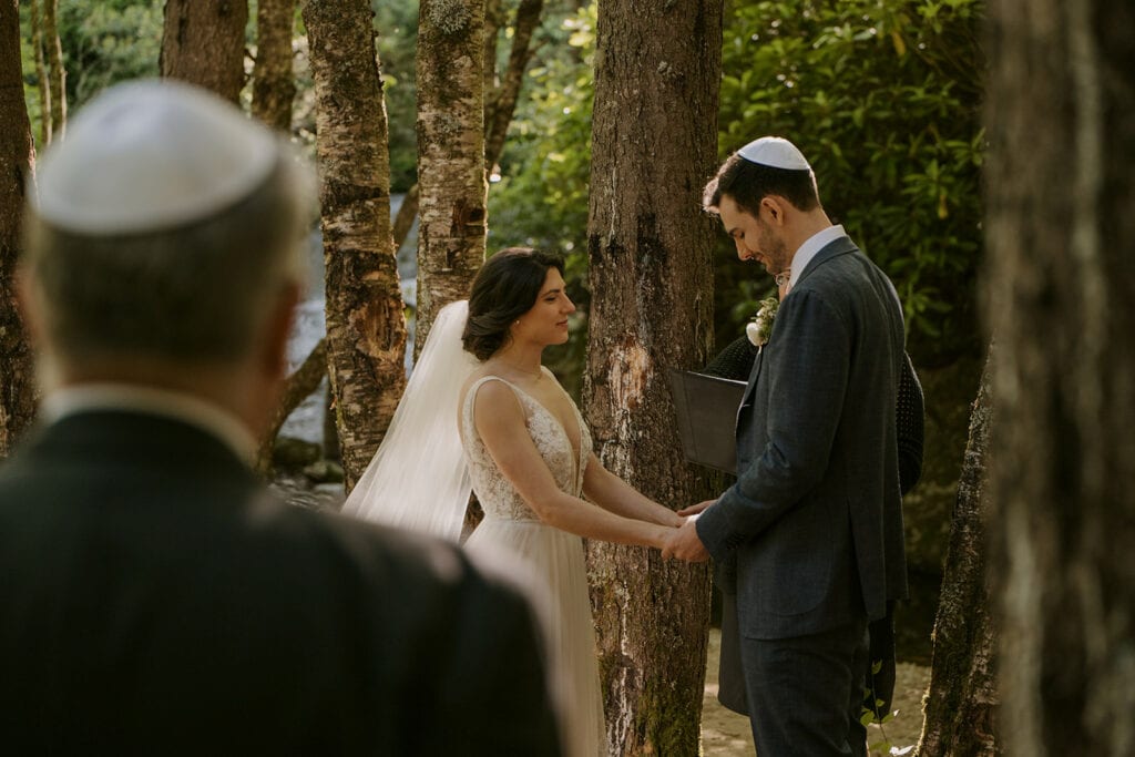 A couple stands holding hands during a Jewish elopement in the forest. A man with a yarmulkes stands in the foreground.