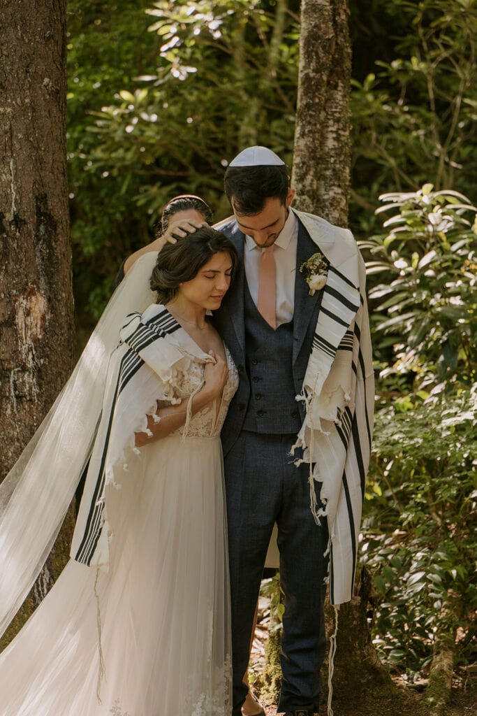 A couple stands side by side with their eyes closed and a Tallit is wrapped around them and their Rabbi prays over them during their Jewish elopement celebration. They are standing in a forest surrounded by trees and rhododendron.