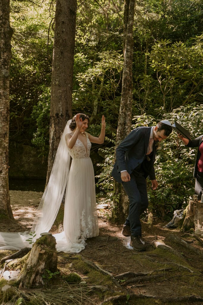 A bride cheers while her husband breaks the glass during their jewish elopement celebration. They are standing in a forest surrounded by trees and rhododendron.