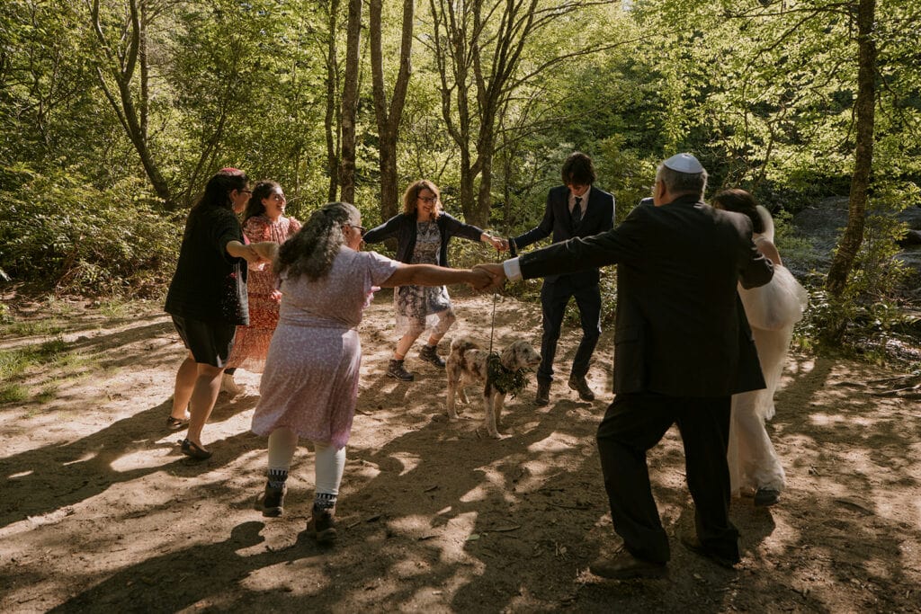 A family dances in a circle during the Hora for a couples Jewish elopement in the forest during summer.