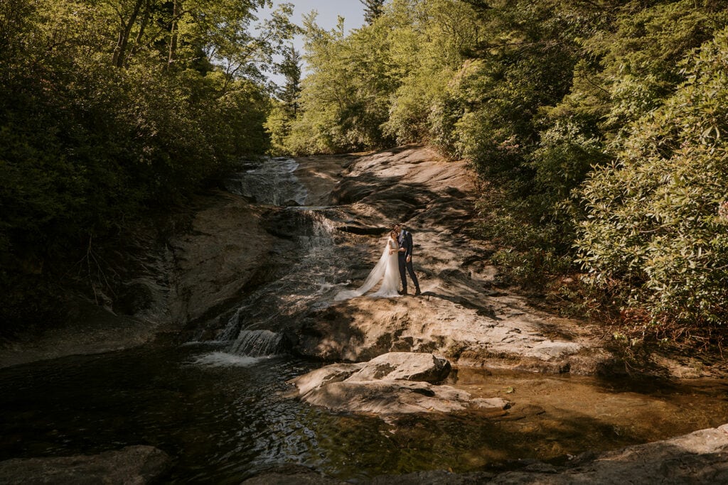 A couple stands side by side smiling and hugging beside a big waterfall in a lush, green forest during their jewish elopement.