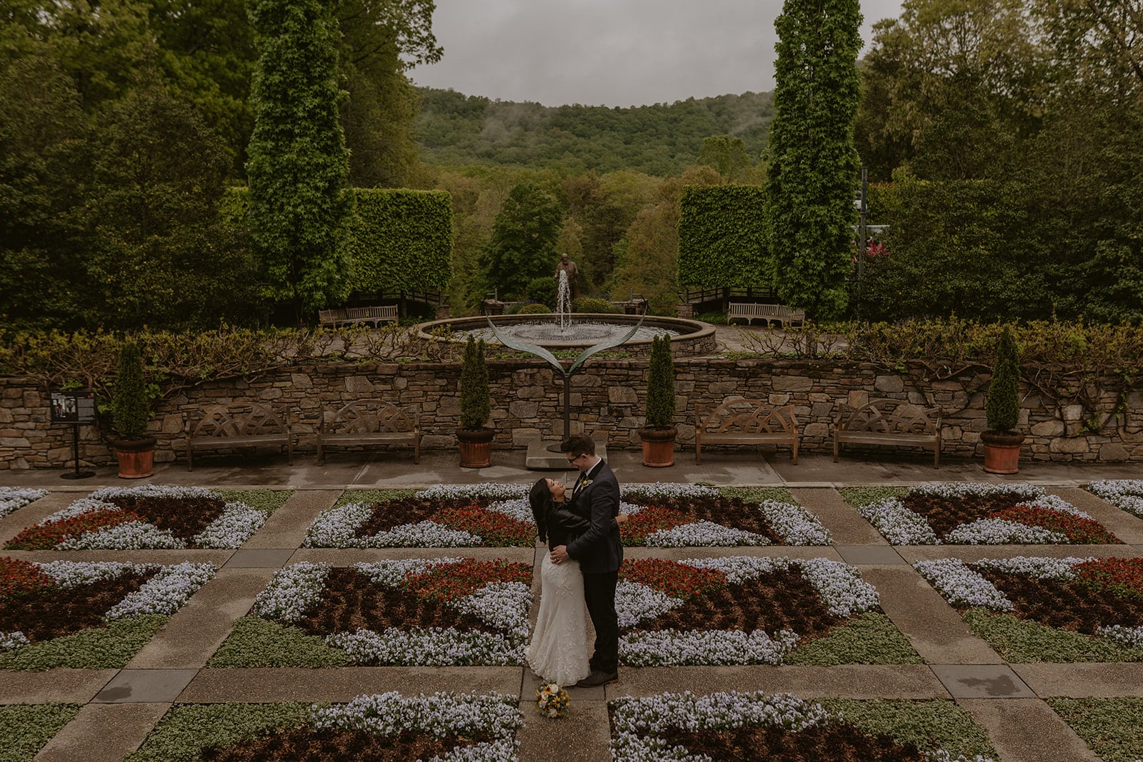 A couple is standing in a blooming field of designed flowers that looks like a quilt. They are in wedding clothes and there is a foggy mountain in the back. They are facing each other smiling with a fountain and square bushes in the background.