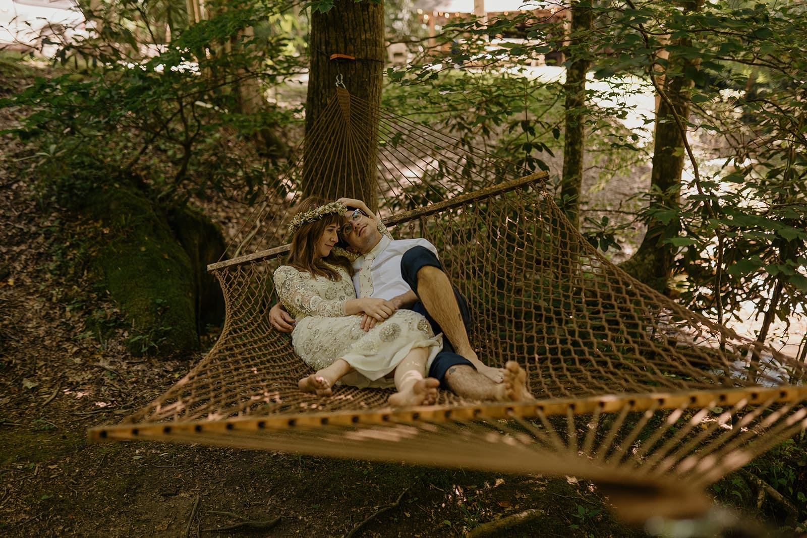 A couple is laying on a hammock for their elopement near Asheville, NC. They are snuggling in their wedding clothes and the groom is looking up at the trees with glasses on while the bride is looking at the river with a flower crown on. They are barefoot.