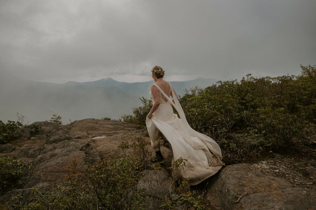 A bride is running toward a mountain view opening up in the clouds with her dress flowing in the wind and hiking boots on for her adventure elopement.