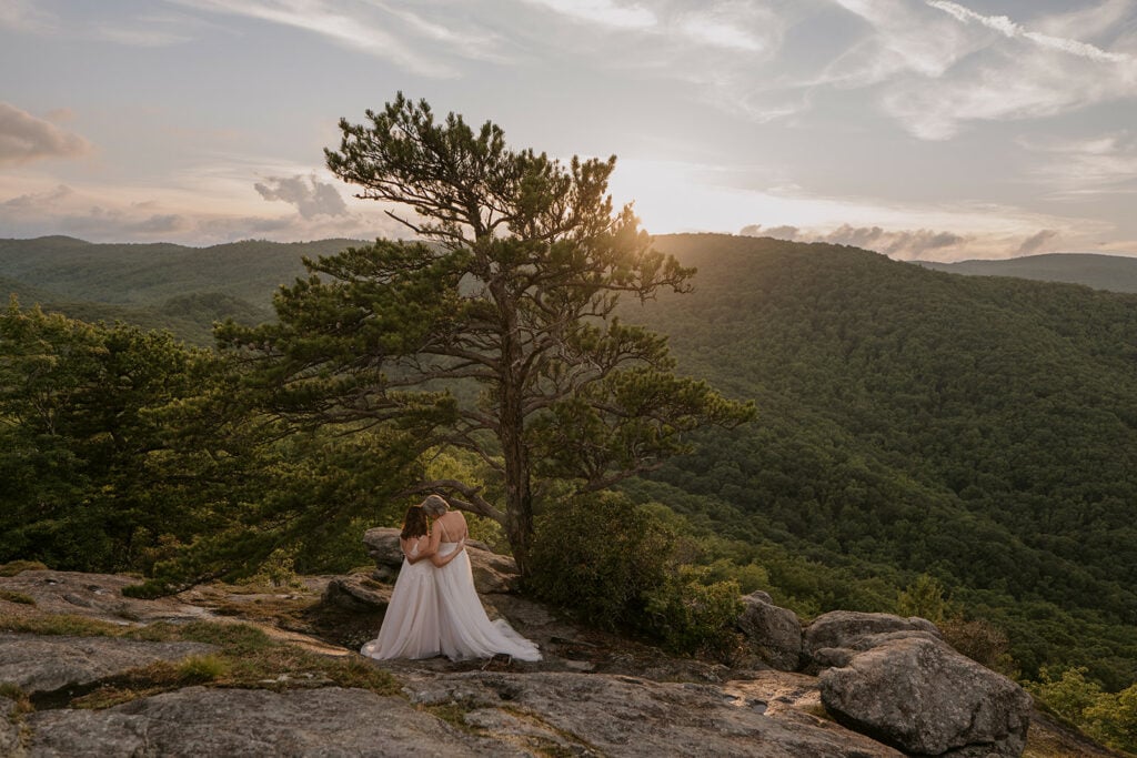 A female couple is standing under a tree at sunset hugging during their elopement near asheville NC. They are both weating long white dresses and have their foreheads pressed together. There is a beautiful mountain view behind them and they are standing on a mossy rock feature.