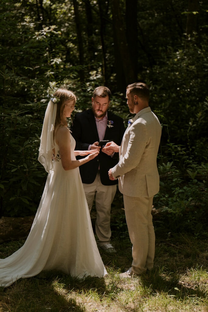 A couple stands in dappled forest light while their officiant hands them rings during a ceremony in North Carolina. They are wearing white and linen outfits.