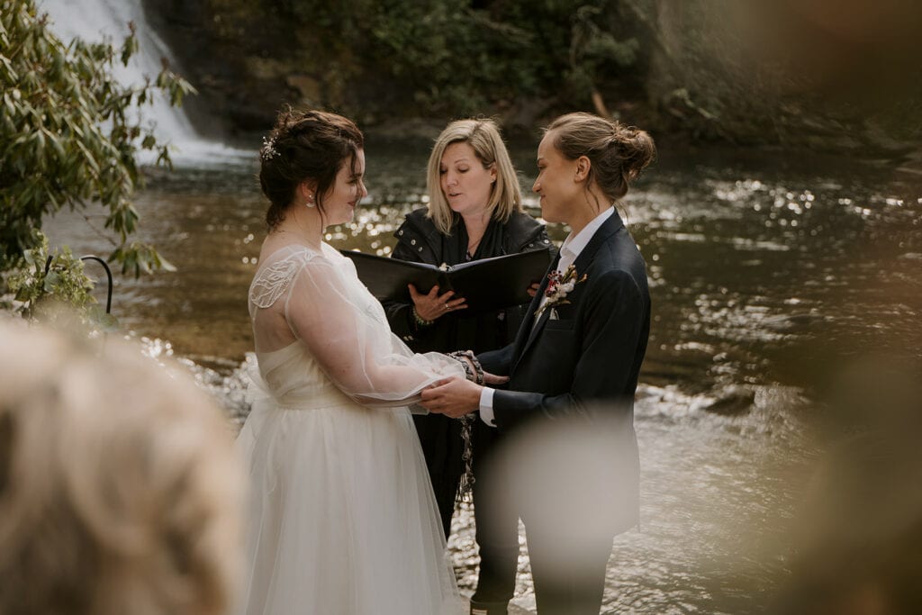 A female couple is standing in front of a waterfall during their wedding in North Carolina while their officiant is reading from their book. They are holding hands and smiling at each other.