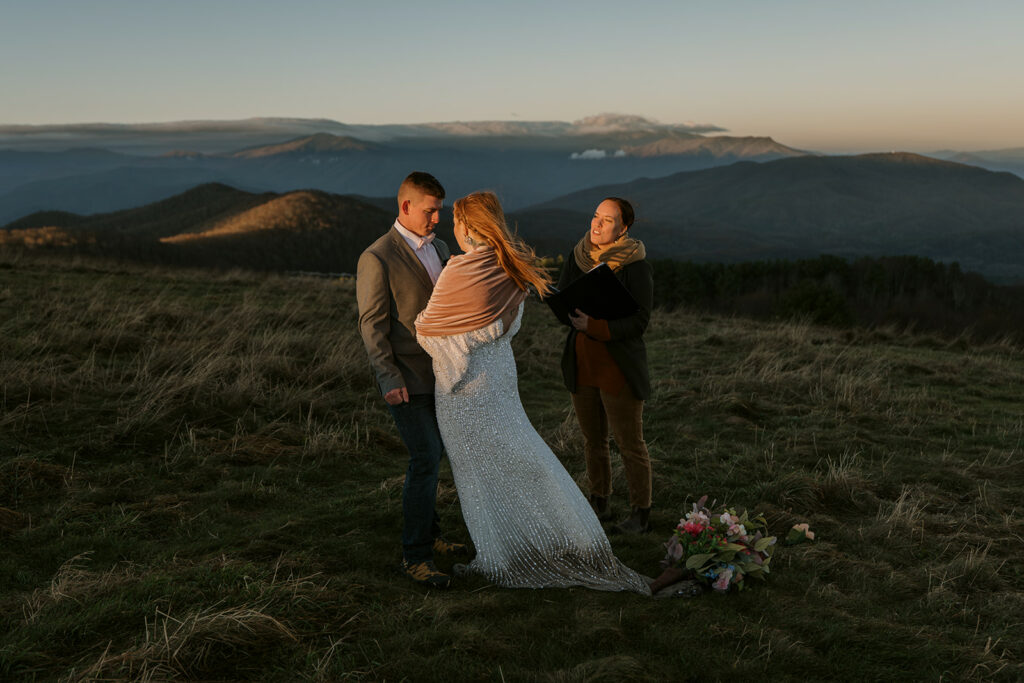 A North Carolina Wedding Officiant reads to a couple at sunrise on a mountain during their wedding. They are lit up in warm sunlight and the mountains are moody in the background.