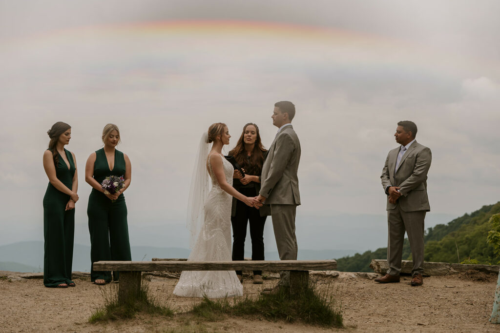 A couple is having a North Carolina Wedding with an officiant reciting words to them while they hold hands. There is a bench, a rainbow, and bridesmaids and groomsmen standing nearby.