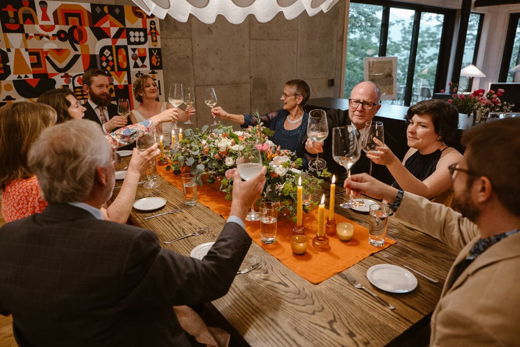 A family is gathered around the table for a wedding dinner party that is intimate. There is a bright orange centerpiece with flowers and candles and they are smiling and cheersing wine glasses.