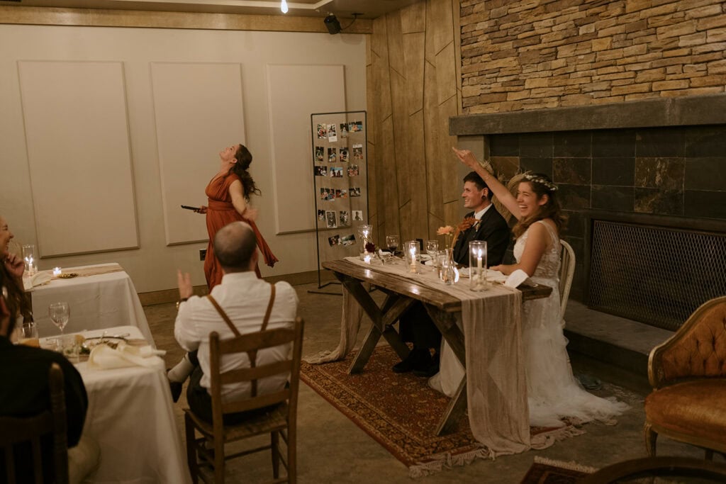 A couple is smiling and laughing and cheering on a friend giving a silly toast during their wedding dinner party. The guests are clapping and they are in a cozy, warm venue.