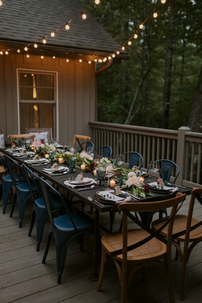 A table set up for a wedding dinner party on a porch with string lights and woods in the back. There are blue and wooden chairs with colorful flowers on a black table with tea lights.
