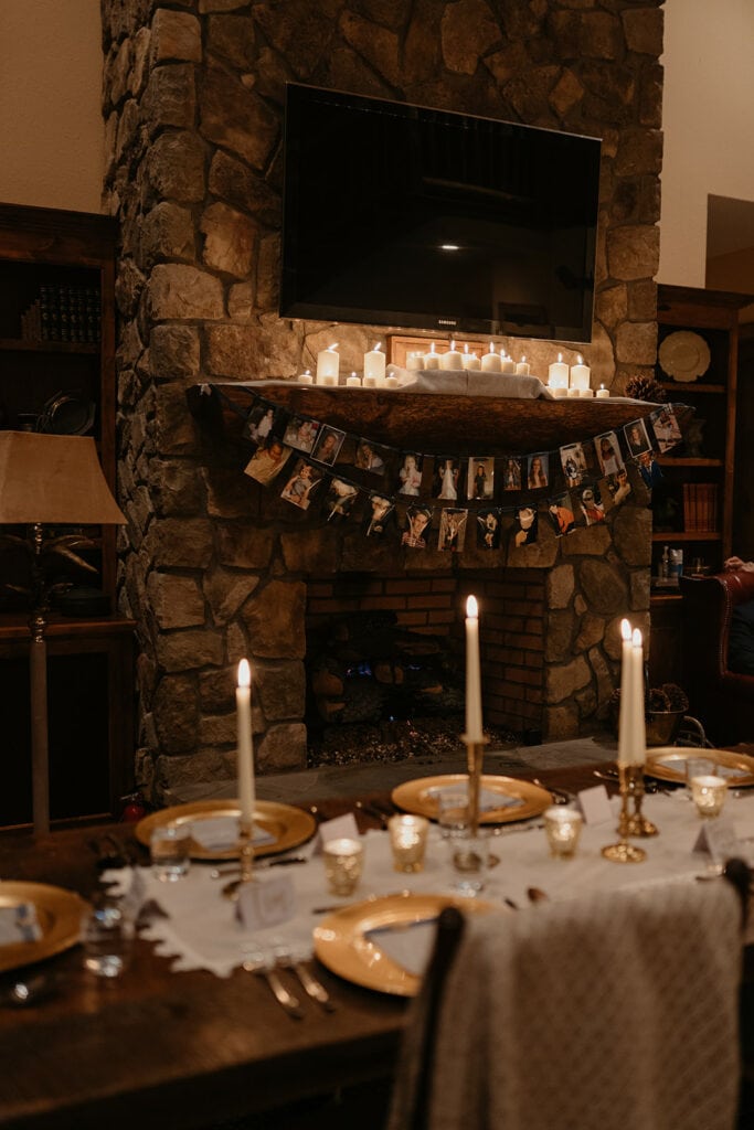 A wedding dinner table is set up in front of a fireplace with candles and hanging pictures. There are candles and golden place settings to set the mood.