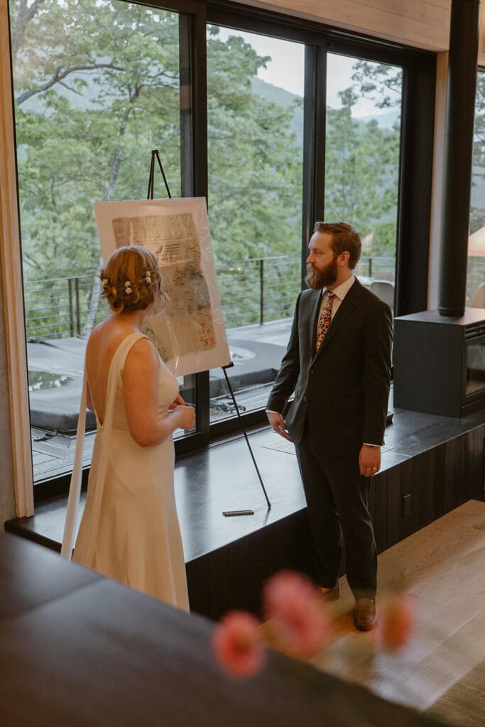 A couple stands in front of a personalized piece of artwork they had a friend create for their wedding dinner party. They are standing and smiling it after everyone has signed it.