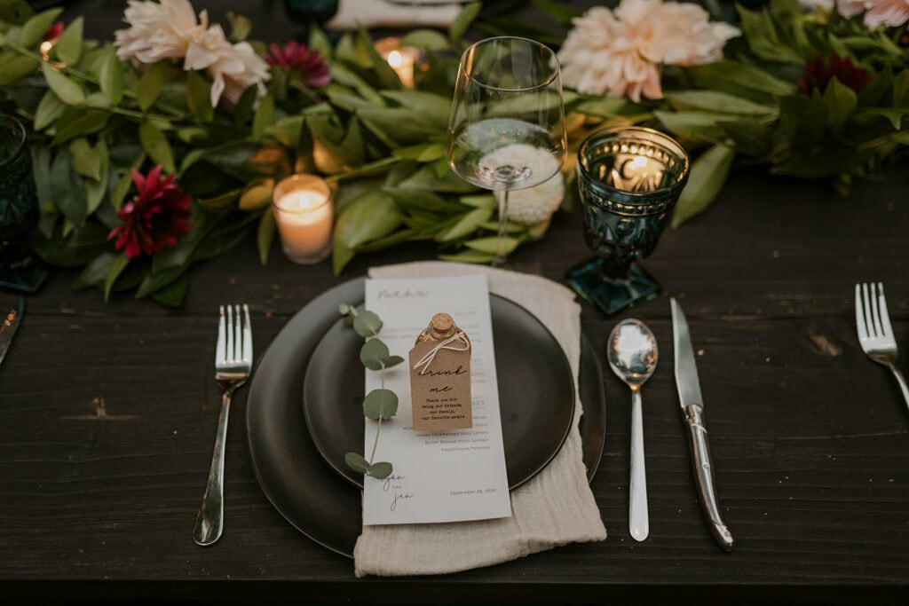 A table setting on a dark wooden table with black plates, a menu, and a bottle that says "drink me" on it. There are flowers and candles for a wedding dinner party.