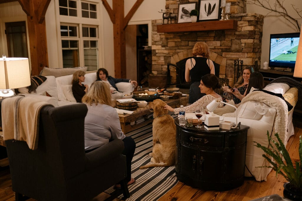 A family is gathered in a cozy living room chatting during a wedding dinner party with food on the coffee table. The bride is sitting and petting a golden retreiver.