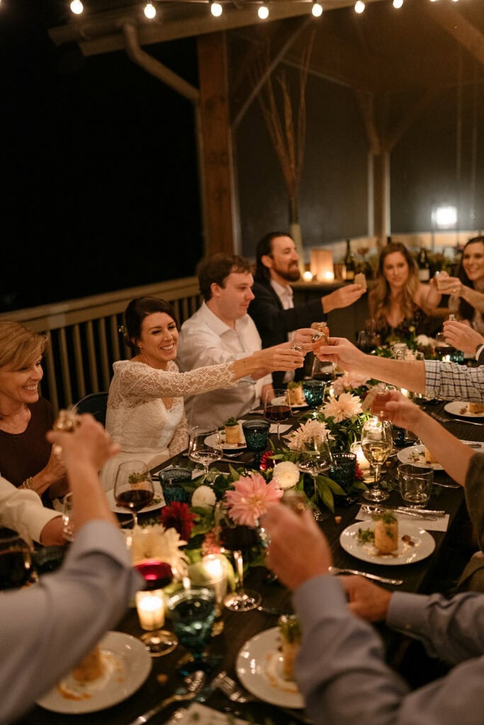 a wedding dinner party around a table lit with candles. They are smiling while they share a cheers with their drinks and the table is covered in good food and flowers.