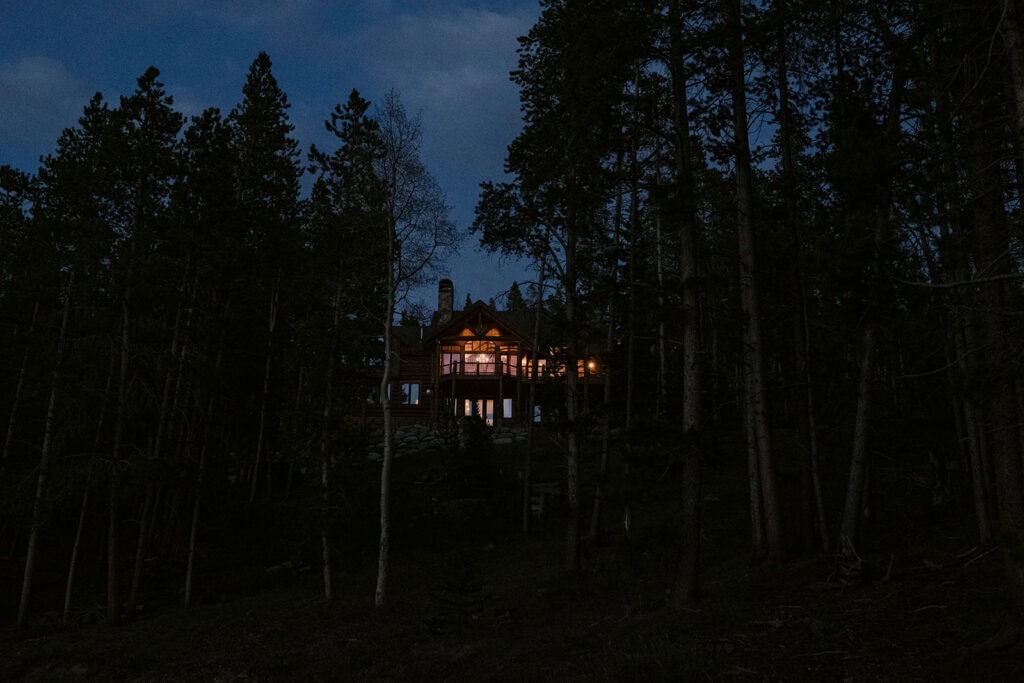A cabin in the woods at dusk is lit up with indoor warm lights. It is a perfect setting for a wedding dinner party.