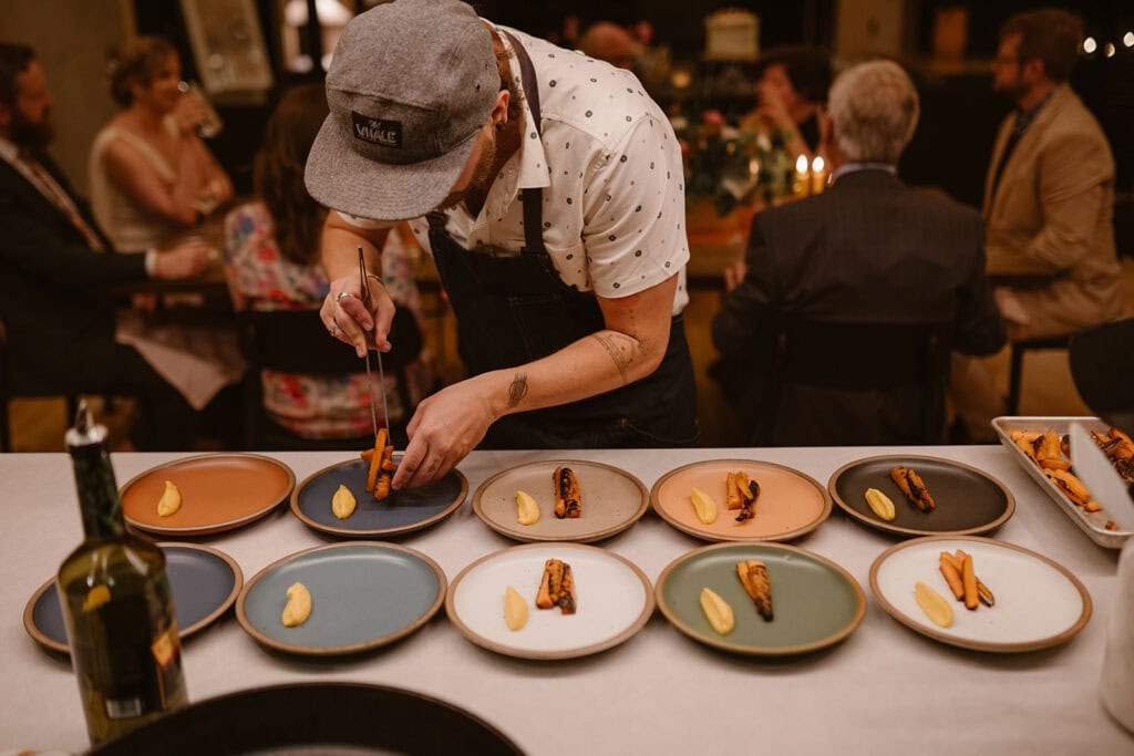 A chef is plating food in front of a group of people sitting at a table for a wedding dinner party.