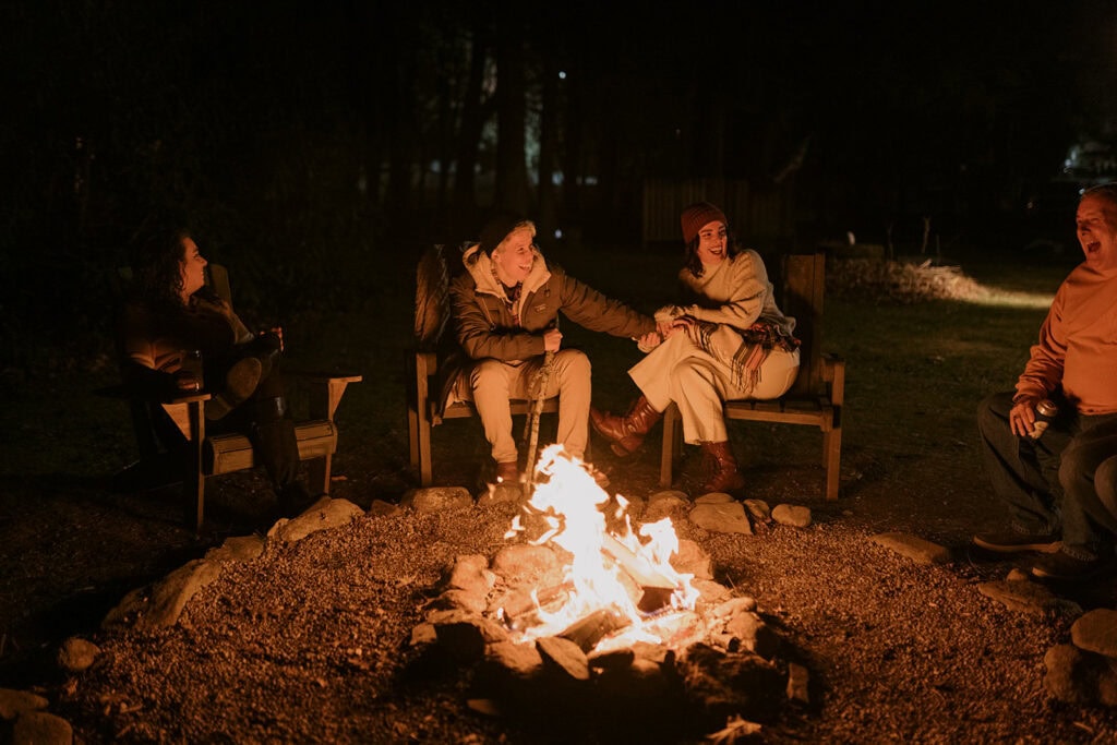 A couple is sitting side by side in front of a fire with their family members after their wedding dinner party. They are laughing and smiling and holding hands.
