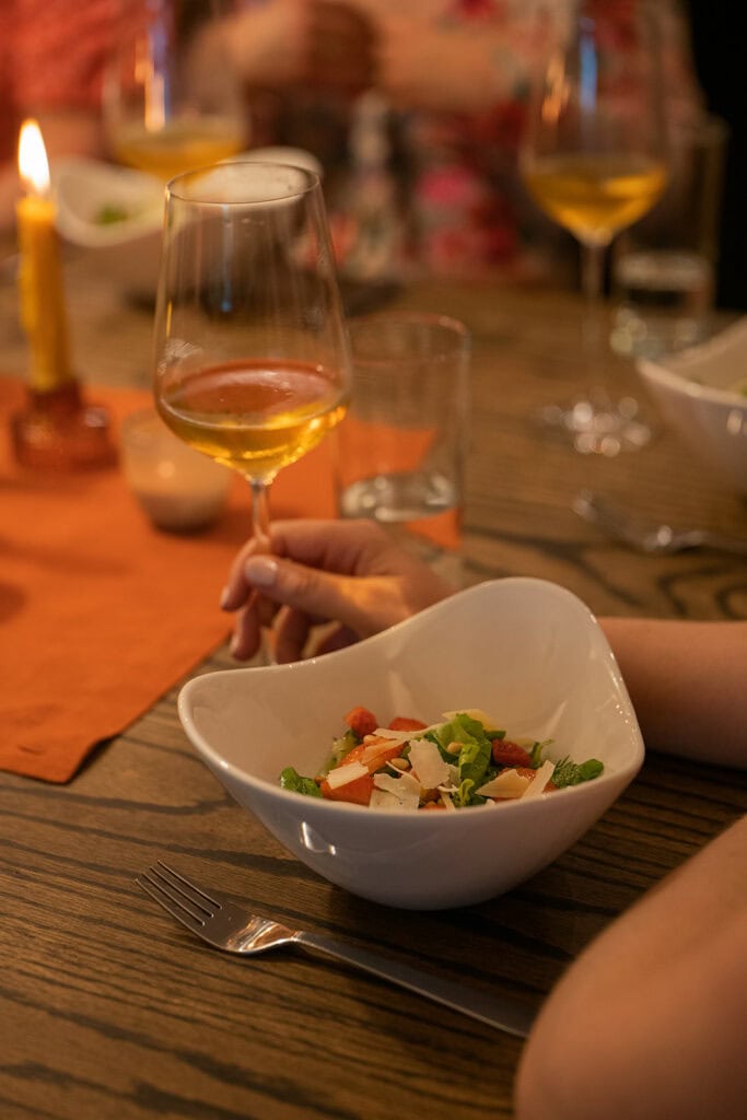 A bowl of fresh salad is sitting in front of a person holding their wine glass during a wedding dinner party. There is warm, glowy light and candles.