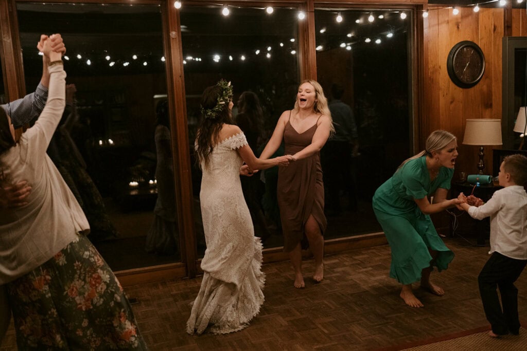 A bride is dancing with her sister in a room filled with string lights and surrounded by family dancing. They are having a blast for their wedding dinner party.