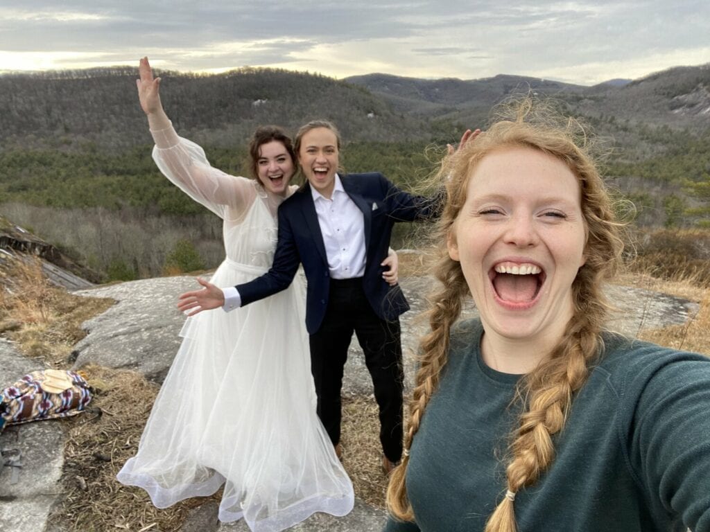A couple and their photographer smile at the camera for a selfie while they elope in Asheville. They are on a mountain in the winter and in wedding clothes with their arms in the air and big smiles on their face. One is in a suit and the other a white dress and the photographer has a big grin with braids.